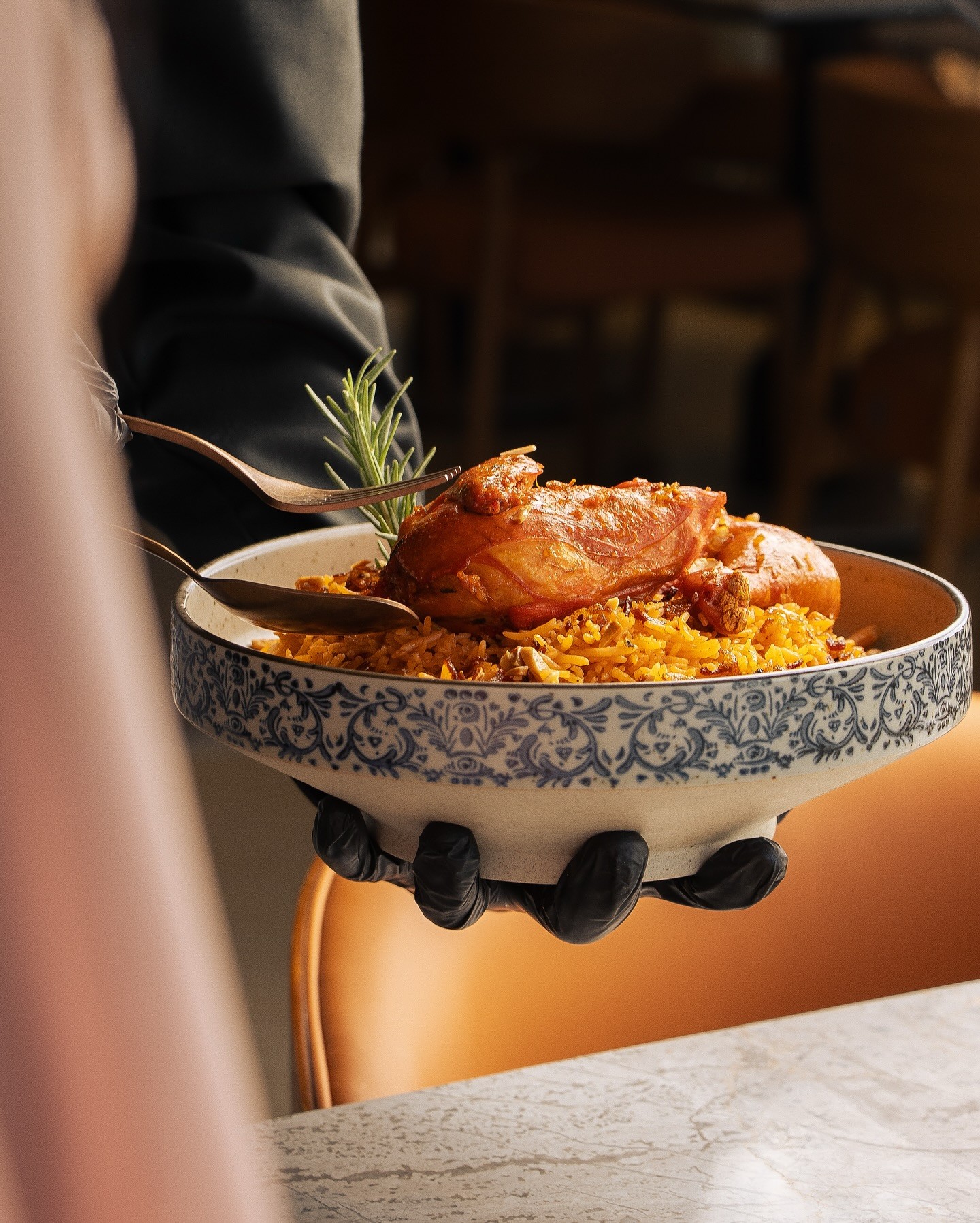 White plate with roasted carrots served on green sauce, topped with almonds and fresh herbs, held by a hand over a dark table.